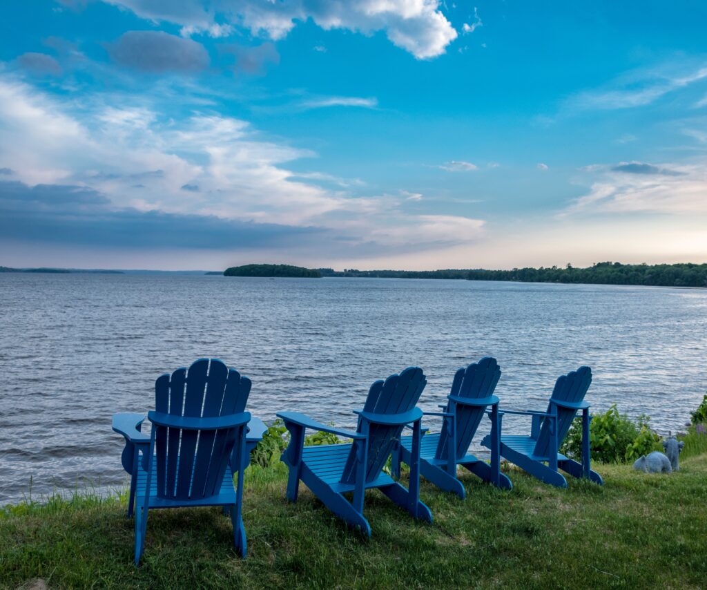 Four adirondack chairs on a grassy shore facing a deep blue lake and partially cloudy sky.