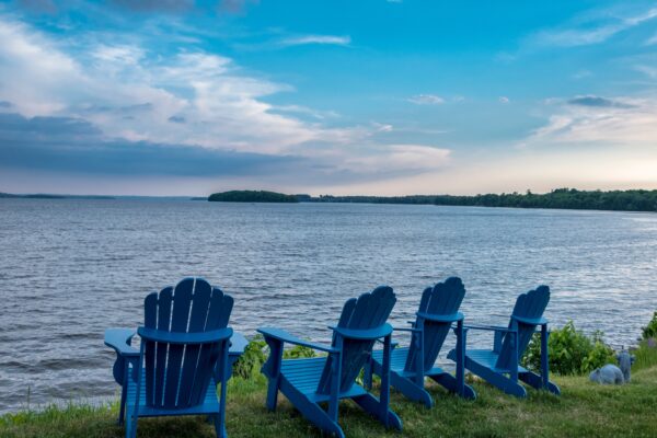 Four adirondack chairs on a grassy shore facing a deep blue lake and partially cloudy sky.